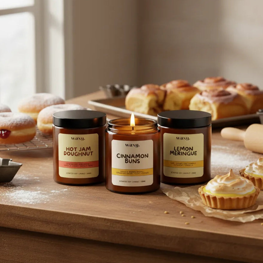 Three candles with labels 'Hot Jam Doughnut', 'Cinnamon Bun', and 'Lemon Meringue' on a wooden surface with pastries in the background.